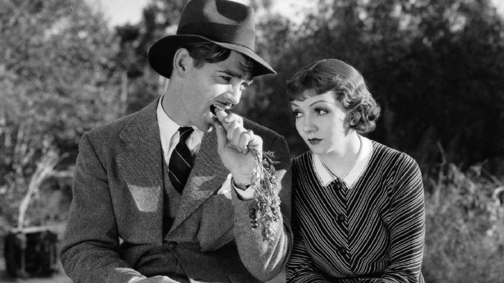 Still from It Happened One Night - Clark Gable chews on a carrot while sitting next to Claudette Colbert on the side of the road, waiting to hitchhike.