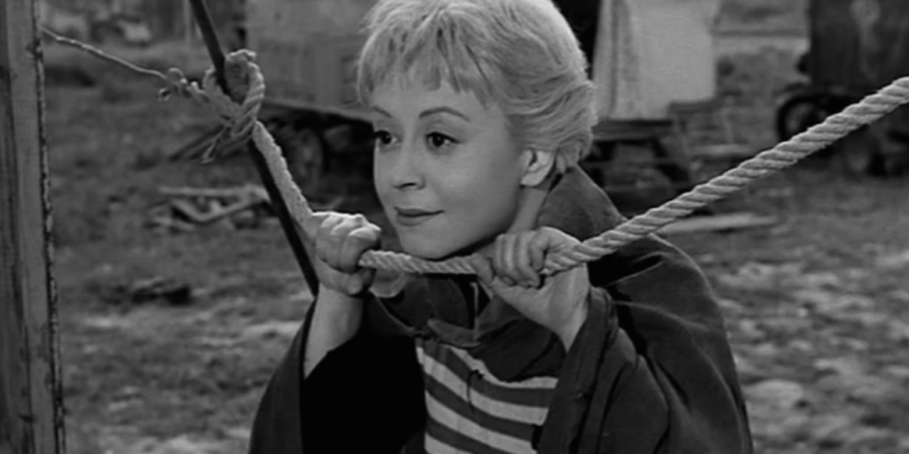 Giulietta Masina as Gelsomina rests her chin on a rope as she watches something on the left of frame.