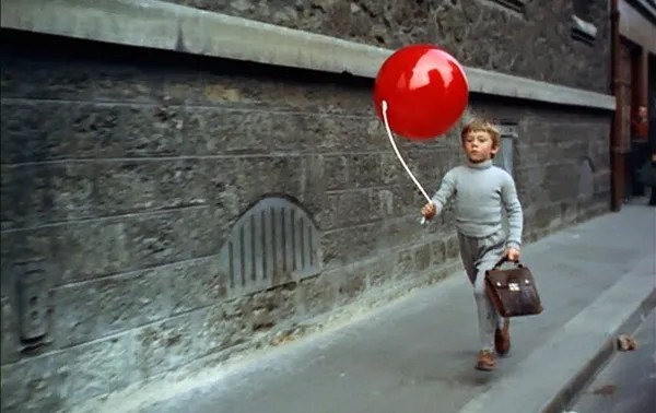 A young boy in gray walks down a gray street, holding the string of a large red balloon.